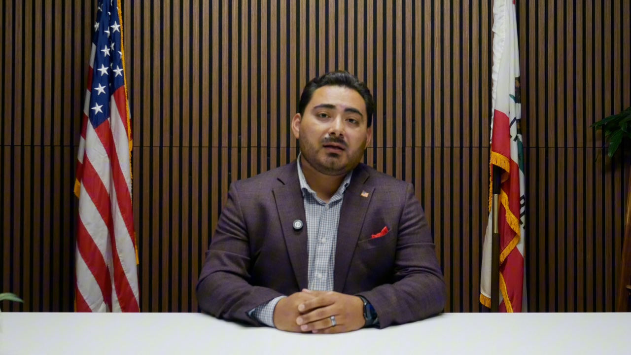 Guillermo Gonzalez speaking to the camera, seated between the American and California flags